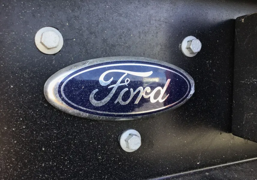 Gros plan sur le logo Ford bleu ovale fixé sur une surface noire légèrement poussiéreuse du véhicule blanc.