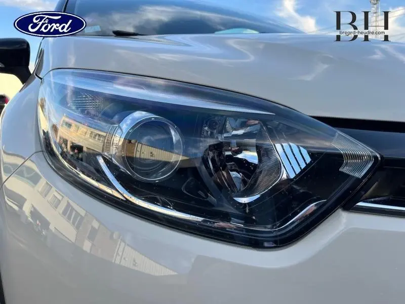 Gros plan sur le phare avant droit d'une Renault Captur beige cendré avec toit noir étoilé, reflet du ciel et bâtiments.