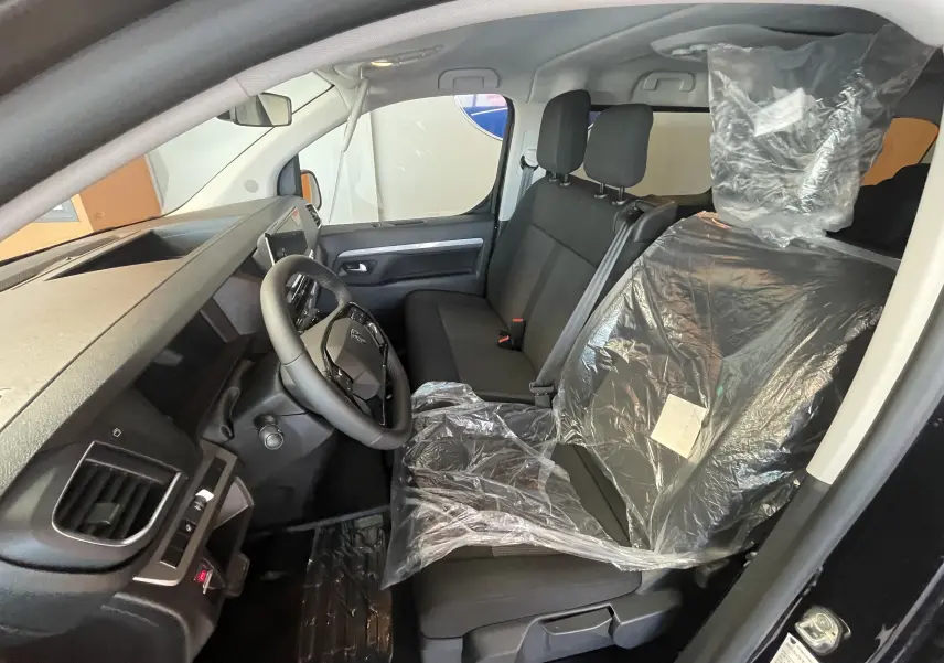 Vue intérieure côté conducteur du Peugeot Traveller noir 2026, montrant le volant cuir et les sièges avant avec protection plastique.