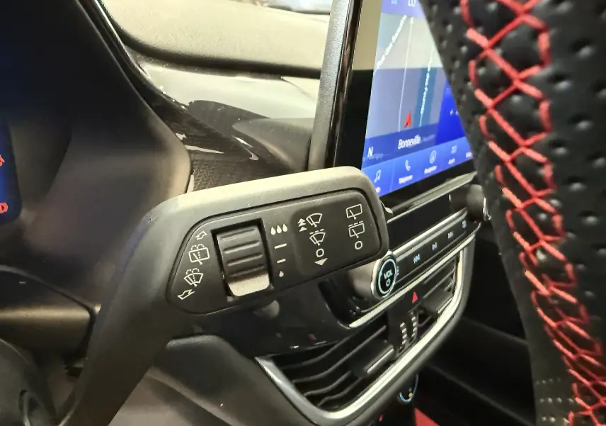 Gros plan sur la commande d’essuie-glace à gauche du volant perforé noir avec surpiqûres rouges, tableau de bord et écran tactile visibles.