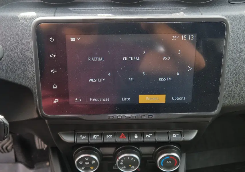 Écran tactile central du Dacia Duster 2022 montrant les stations radio, avec commandes climatisation et boutons fonction sous l'écran.