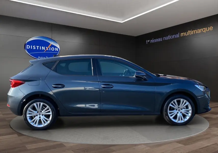 Profil droit d'une SEAT Leon IV gris magnetic avec jantes alu 17 pouces, dans un showroom moderne.