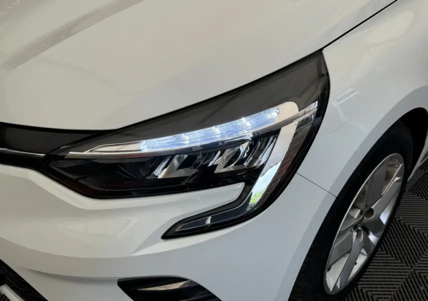 Gros plan sur le phare avant droit allumé d'une Renault Clio Business blanche, avec jante alu visible.