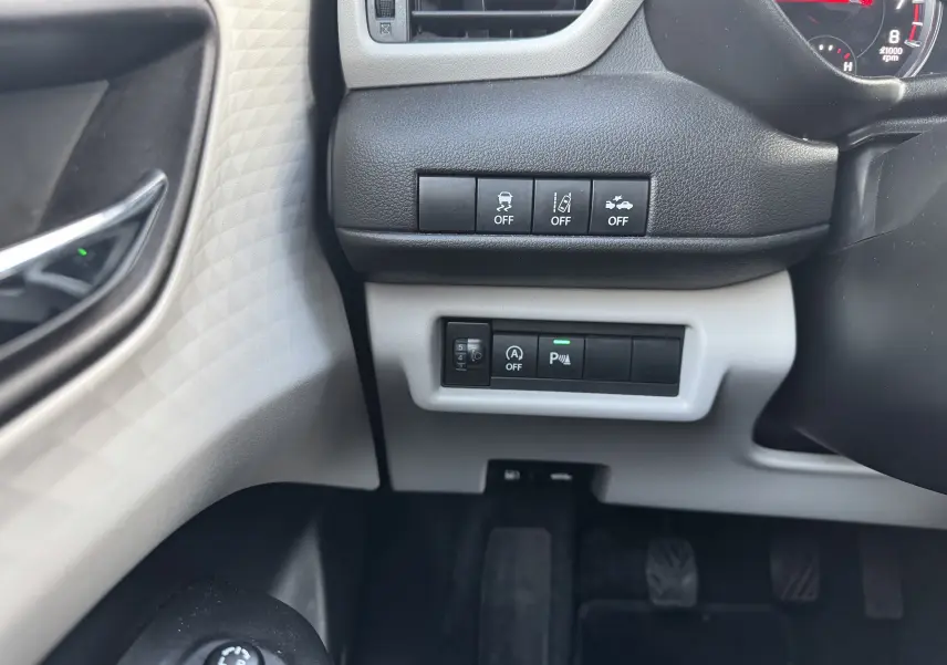 Détail des commandes sous le tableau de bord côté gauche d'une Suzuki Swift blanche, avec boutons d'assistance et réglages lumineux.