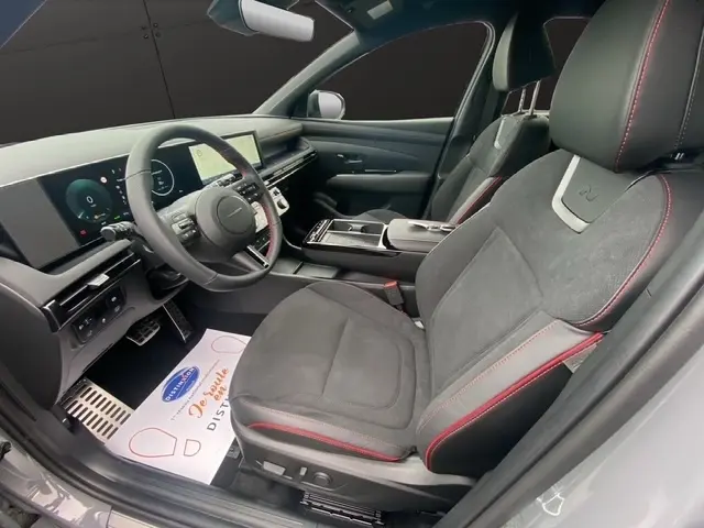 Vue intérieure côté conducteur du Hyundai Tucson 1.6 Hybrid gris foncé, sièges en alcantara avec surpiqûres rouges et tableau de bord numérique.