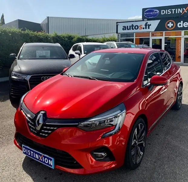 Renault Clio V rouge vue 3/4 avant droit, avec calandre distinctive et jantes alliage, stationnée devant un concessionnaire.