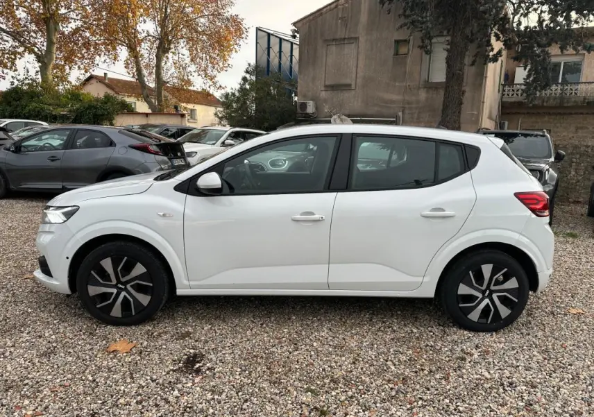 Profil côté gauche d'une Dacia Sandero blanche 2024 garée sur un sol gravillonné, avec jantes noires et grises.