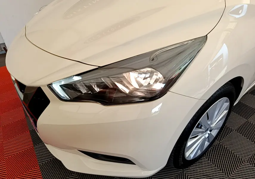 Vue 3/4 avant droit d'une Nissan Micra IG-T 92 Acenta blanche avec phares allumés et jantes alliage visibles.