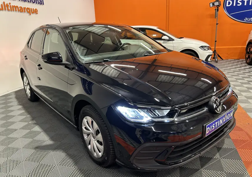 Volkswagen Polo noire vue en 3/4 avant droit avec phares allumés dans un showroom orange.