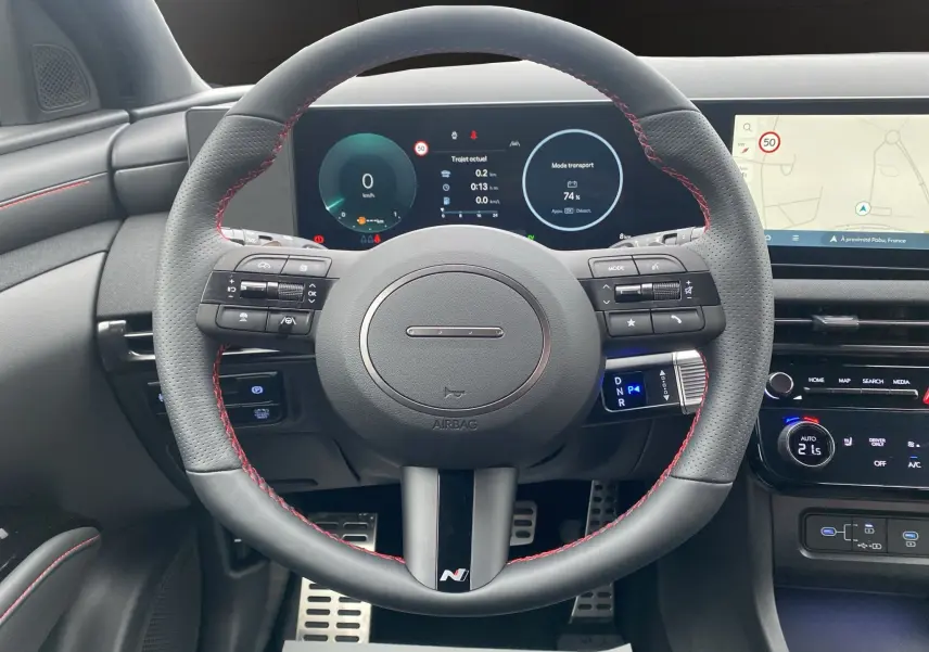 Vue frontale du volant cuir noir avec surpiqûres rouges du Hyundai Tucson 2026, tableau de bord digital et écran GPS visibles.