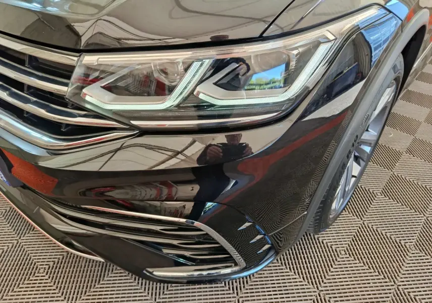 Gros plan sur le phare avant droit noir brillant du Volkswagen Tiguan 1.4 eHybrid 2023 avec détails chromés et jante visible.
