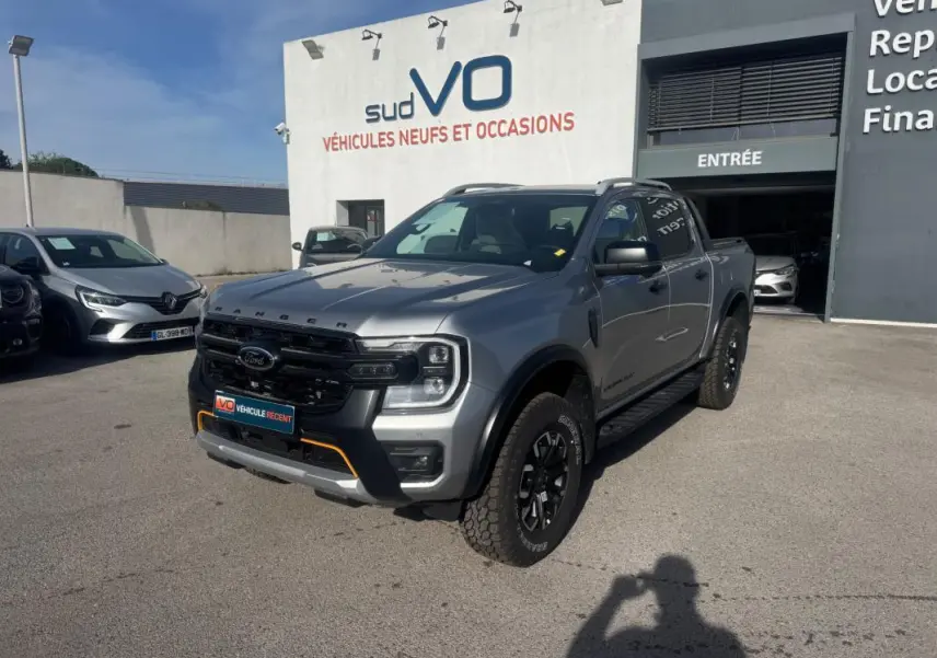 Ford Ranger Wildtrak gris en 3/4 avant droit, avec barres de toit et jantes alliage noires visibles.