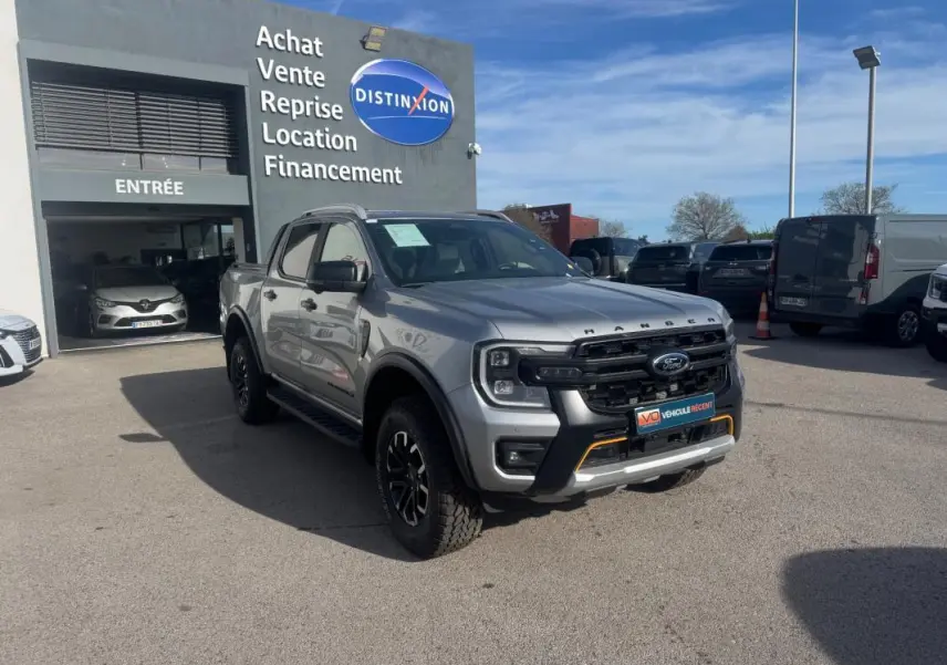 Ford Ranger gris en 3/4 avant droit, avec jantes alliage noires et barres de toit visibles, stationné devant un garage.