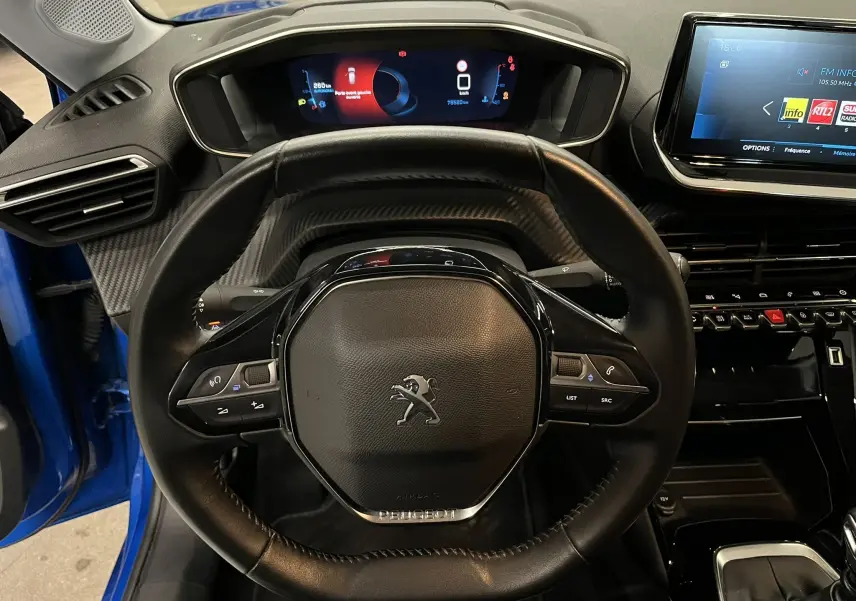 Vue intérieure centrée sur le volant compact de la Peugeot 208 bleu Vertigo, avec tableau de bord numérique et écran tactile visible.