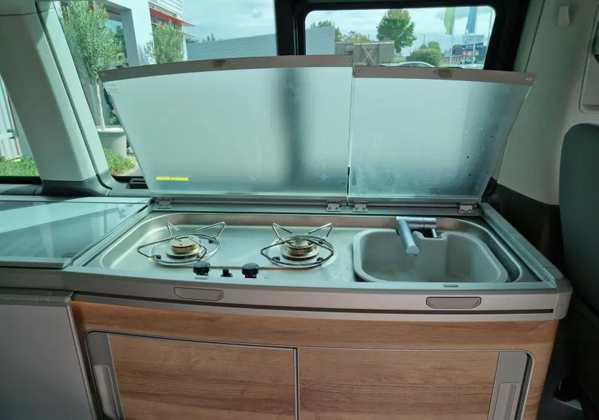 Cuisine intégrée avec deux feux gaz et évier dans un Volkswagen California Cooper bronze, vue intérieure côté droit.