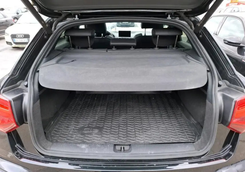 Coffre ouvert vu de l'arrière d'un Audi Q2 noir avec cache-bagages et tapis de coffre en caoutchouc.