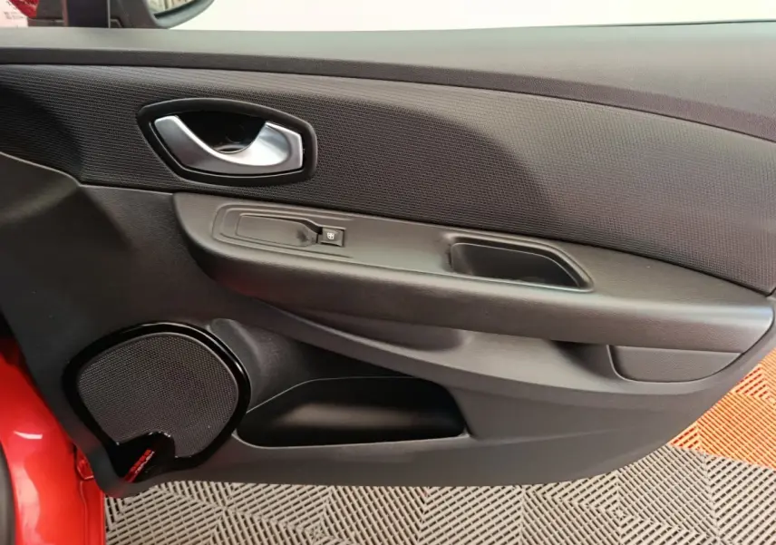 Détail de la porte intérieure côté conducteur de la Renault Clio rouge, avec poignée argentée et haut-parleur intégré.