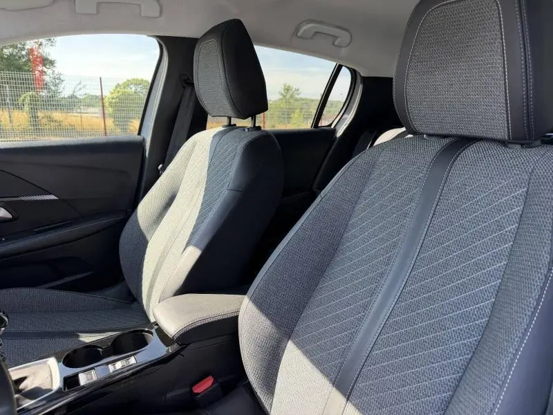 Vue intérieure de profil montrant les sièges avant en tissu noir et gris avec surpiqûres blanches de la Peugeot 208 2025.