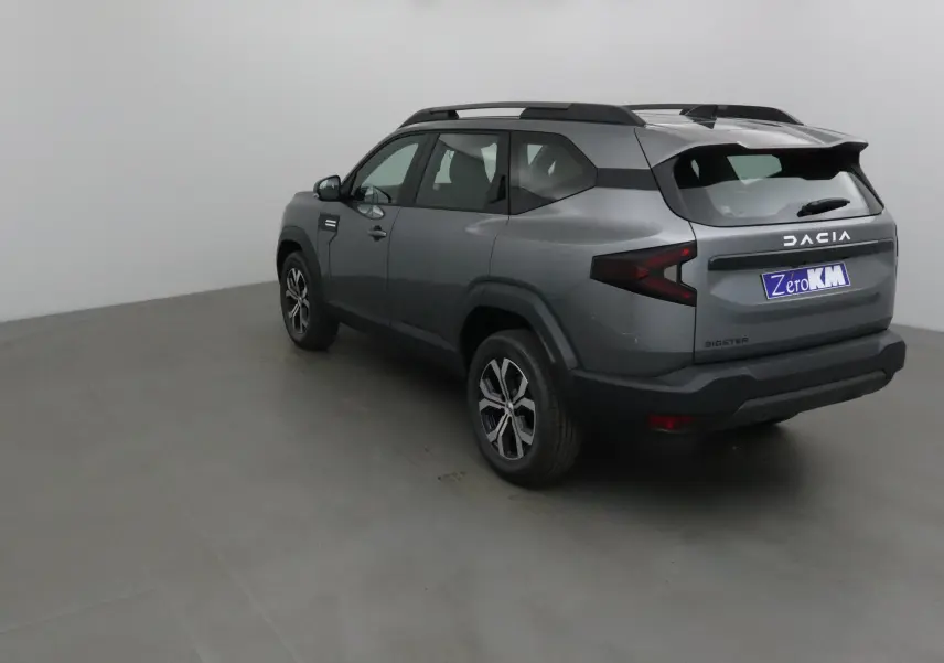 Vue 3/4 arrière droite du Dacia Bigster gris schiste 2025 avec feux arrière en forme de flèche et barres de toit noires.