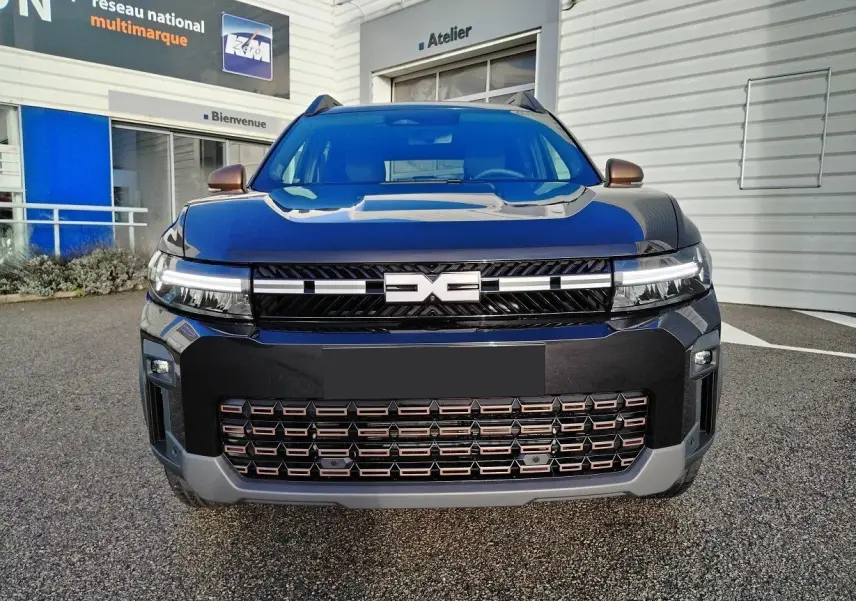 Vue frontale d'un Dacia Bigster noir 2025 avec calandre ajourée et phares LED fins, stationné devant un atelier.