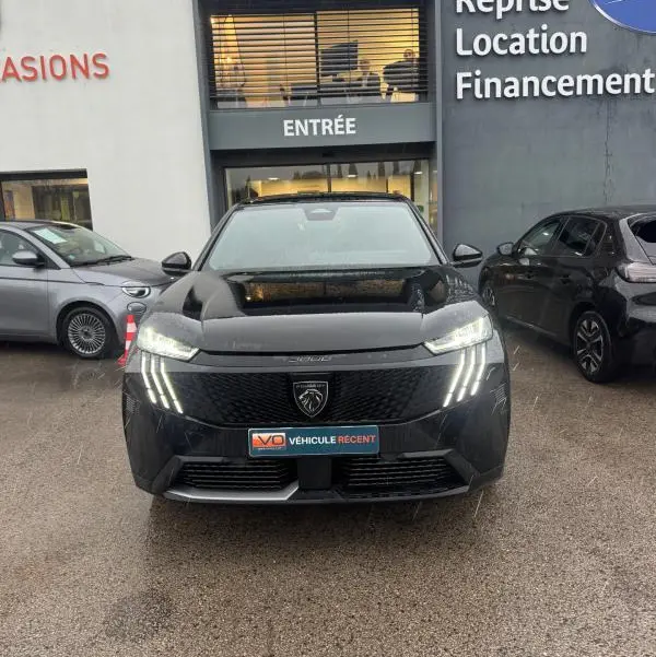 Vue de face du Peugeot 3008 Hybrid noir avec feux diurnes LED en forme de griffes, garé devant un bâtiment commercial.