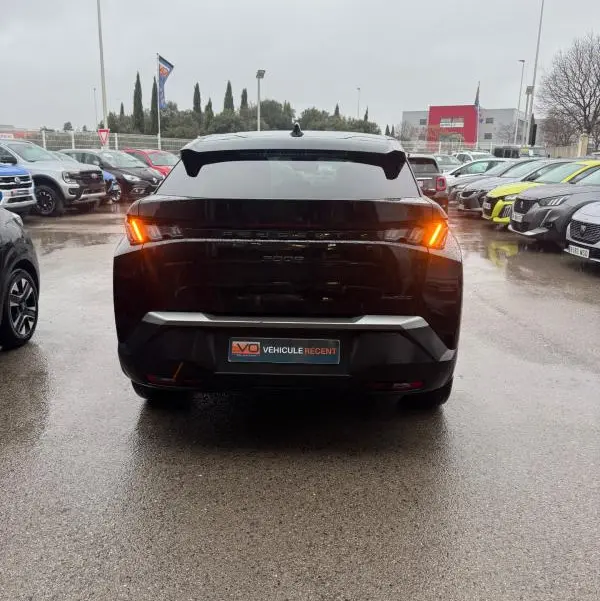 Vue arrière d'un Peugeot 3008 Hybrid 145 noir avec feux allumés, garé sur un parking humide entouré d'autres véhicules.