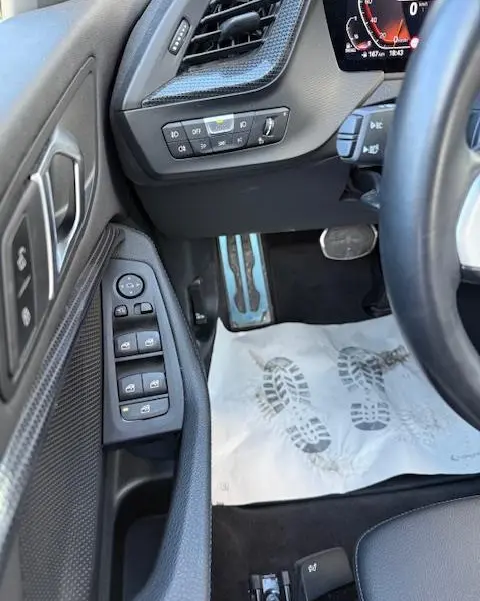 Vue intérieure côté conducteur de la BMW Série 1 118d Edition Sport 2024, montrant les commandes de porte et pédales en aluminium.