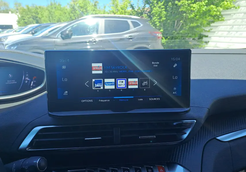 Vue intérieure du tableau de bord du Peugeot 3008 2023, écran central tactile affichant les radios FM, avec extérieur et voiture grise en arrière-plan.