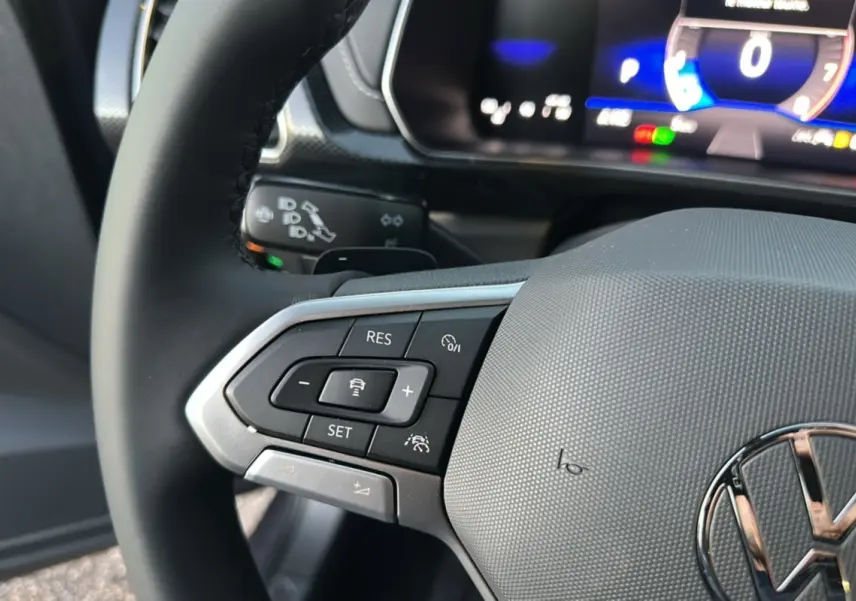 Gros plan sur le volant cuir noir du Volkswagen T-Cross gris cendré, avec commandes multifonctions et tableau de bord numérique en arrière-plan.