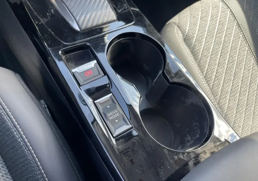 Gros plan sur la console centrale noire brillante avec boutons frein à main électrique et mode de conduite, siège tissu gris à côté.