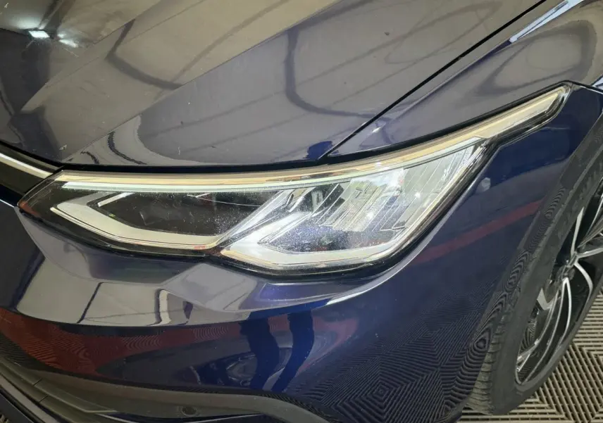Gros plan sur le phare avant droit d'une Volkswagen Golf bleu foncé, avec une jante noire visible en arrière-plan.