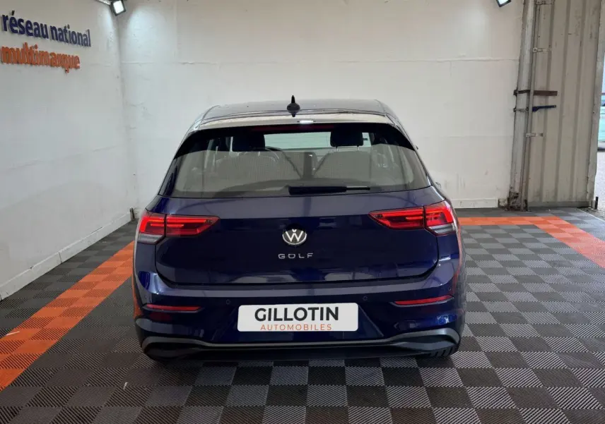 Vue arrière d'une Volkswagen Golf bleu foncé garée en intérieur sur sol à damiers gris et orange.