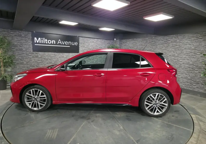 Profil droit d'une KIA RIO rouge métallisée 2022 avec jantes alliage 17'' et vitres surteintées en intérieur showroom.