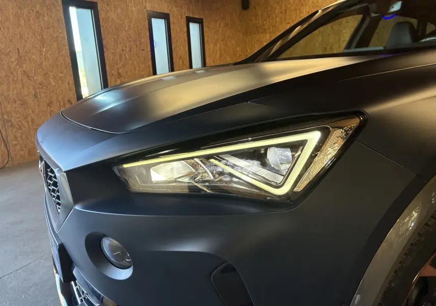Gros plan sur le phare avant droit allumé d'une CUPRA Formentor 1.4 e-Hybrid 2023 bleu mat, avec calandre et capot visibles.