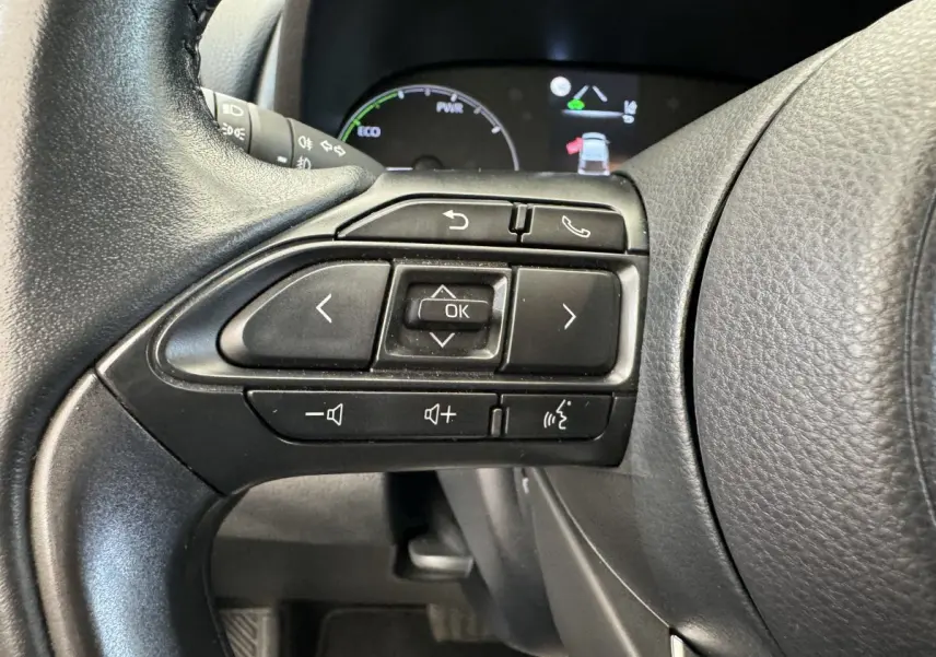 Gros plan sur les commandes multifonctions du volant cuir noir de la Toyota Yaris hybride blanche, tableau de bord en arrière-plan.