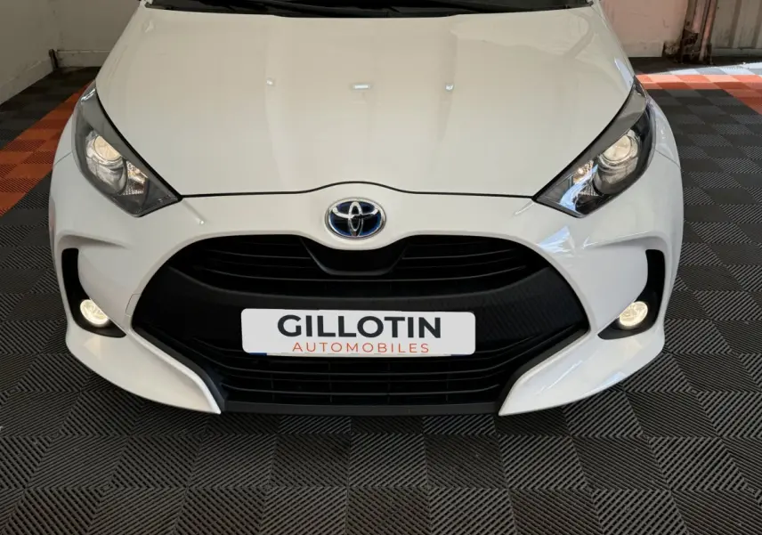 Vue frontale rapprochée d'une Toyota Yaris blanche avec calandre noire et phares allumés dans un showroom.