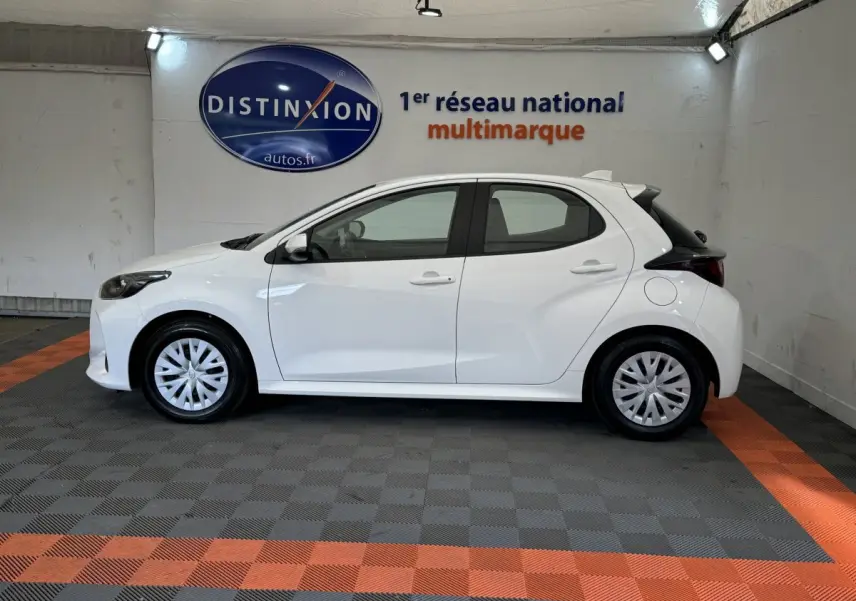 Profil côté gauche d'une Toyota Yaris hybride blanche 2022, dans un showroom avec sol gris et bandes orange.