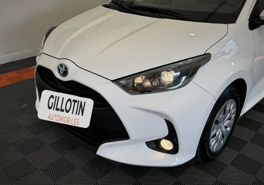 Vue 3/4 avant droit d'une Toyota Yaris blanche avec phares allumés et calandre noire distinctive.