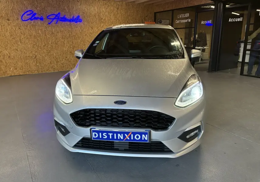 Vue avant d'une Ford Fiesta 1.0 EcoBoost gris avec phares allumés dans un showroom moderne.