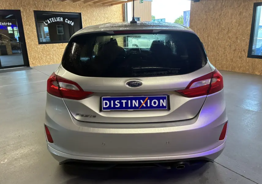 Vue arrière d'une Ford Fiesta gris clair 2020 avec feux arrière rouges et plaque Distinxion en intérieur showroom.