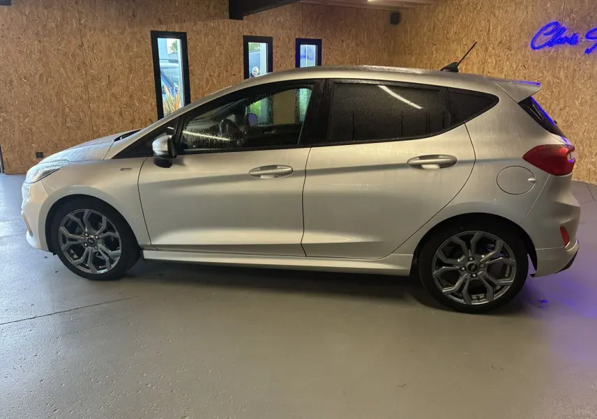 Ford Fiesta 1.0 EcoBoost ST Line gris vue de profil côté gauche dans un intérieur avec sol gris et mur en bois.
