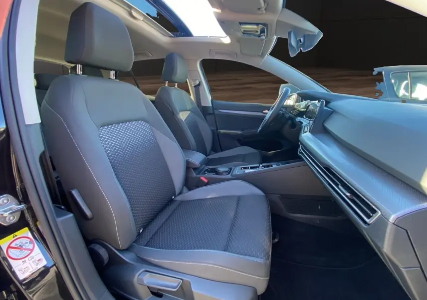 Vue intérieure côté gauche de la Volkswagen Golf VIII noire, mettant en valeur les sièges tissu noirs et le toit panoramique.