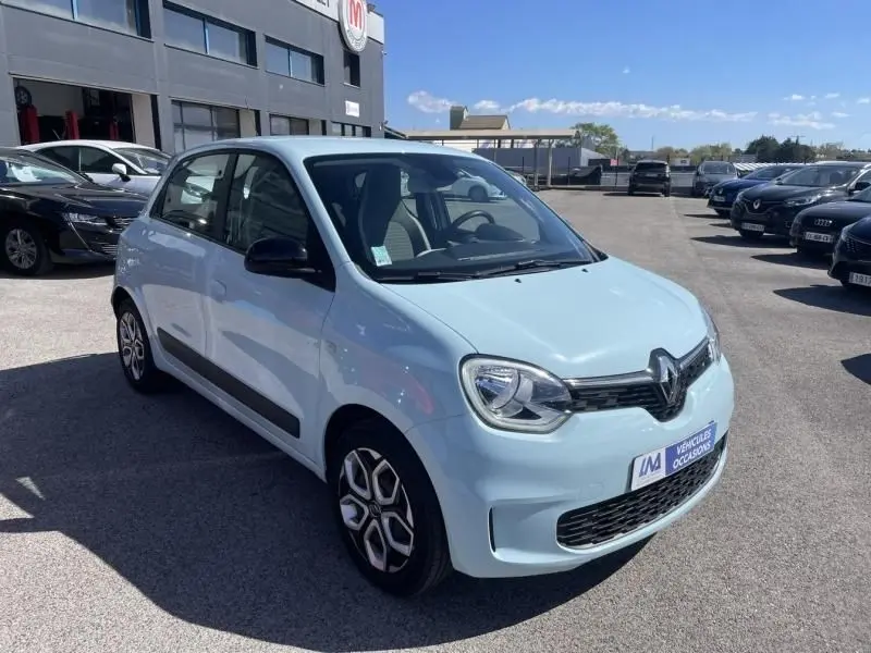 Renault Twingo III bleu dragée vue 3/4 avant droit sur parking, avec jantes distinctives et rétroviseurs noirs.