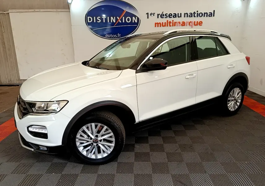 Vue 3/4 avant droite du Volkswagen T-ROC blanc avec jantes alliage et barres de toit noires en intérieur showroom.