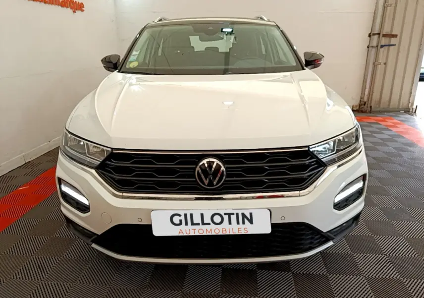 Vue frontale d'un Volkswagen T-Roc blanc avec calandre noire et feux LED allumés en intérieur showroom.