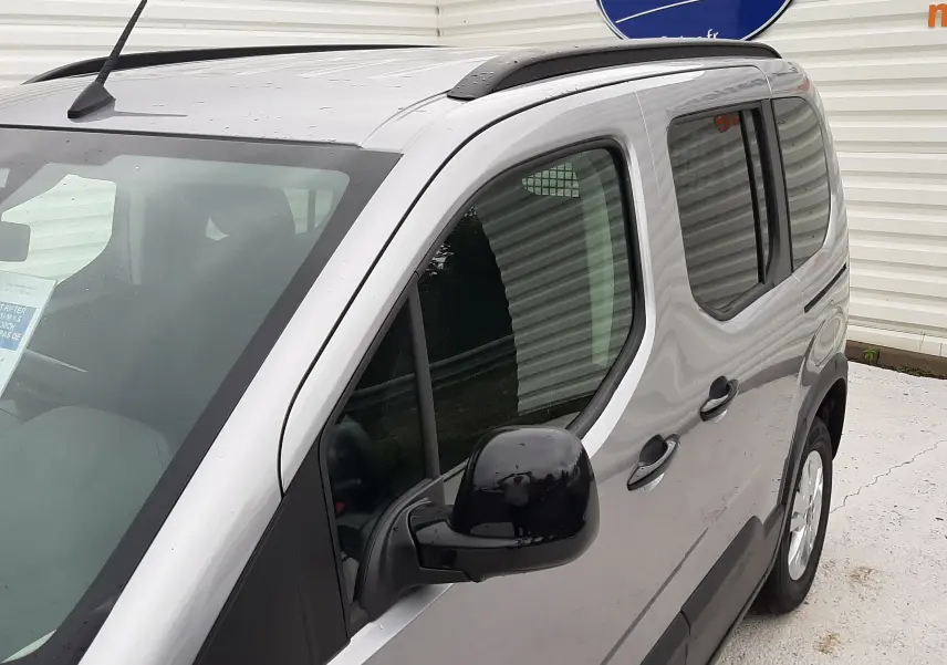 Vue 3/4 avant droit du Peugeot Rifter gris acier avec barres de toit noires et vitres arrière surteintées.
