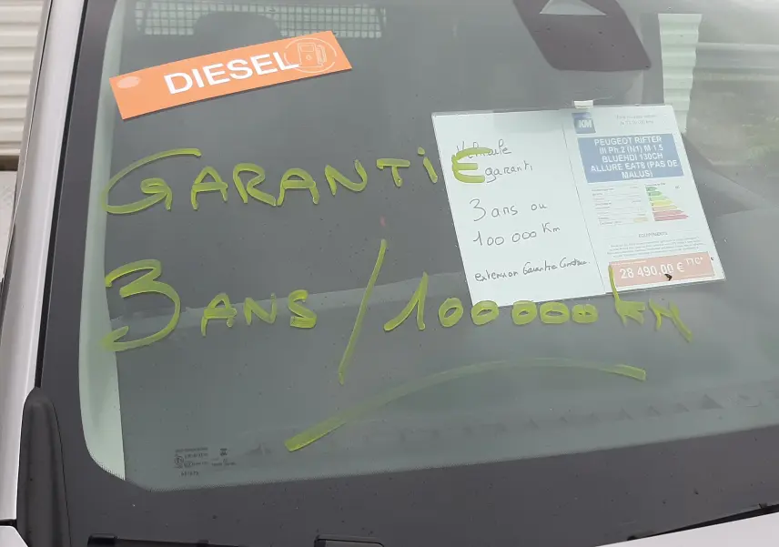 Vue avant rapprochée du Peugeot Rifter gris acier avec inscriptions de garantie et étiquette diesel sur le pare-brise.