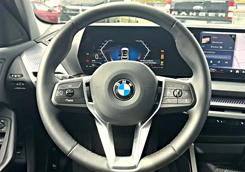 Vue intérieure centrée sur le volant cuir noir BMW d'une Série 1 120da 2025 avec tableau de bord numérique et écran tactile.