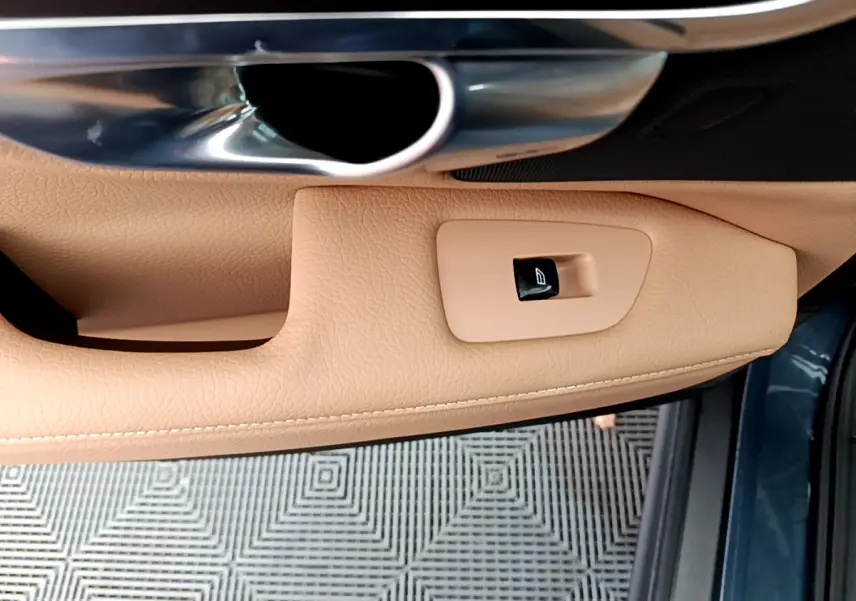 Détail de la commande de lève-vitre sur la porte côté conducteur en cuir beige, avec poignée métal et bord bleu extérieur visible.