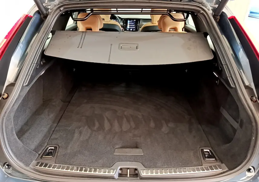 Coffre ouvert vu de l'arrière d'un Volvo V90 Cross Country bleu, avec cache-bagages et intérieur beige visible.